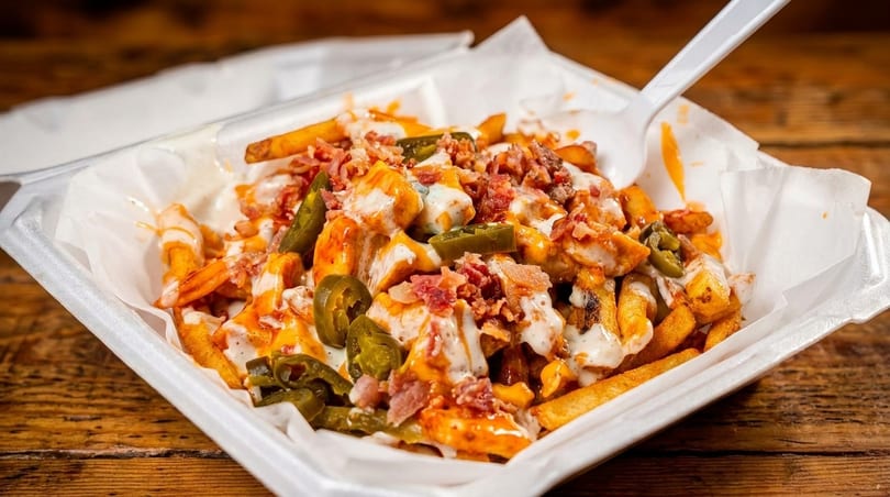 Loaded fries with chicken, bacon, jalapeños, and white sauce in a takeout container.