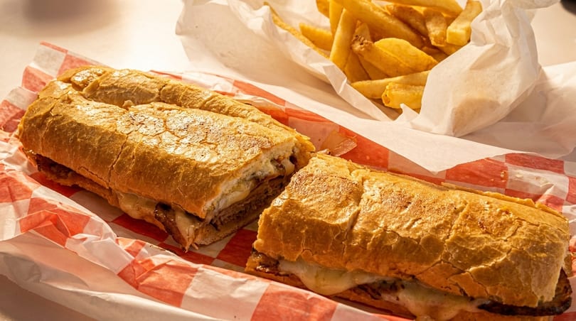 A sliced cheesesteak sandwich with melted cheese and a side of golden fries in paper-lined baskets.