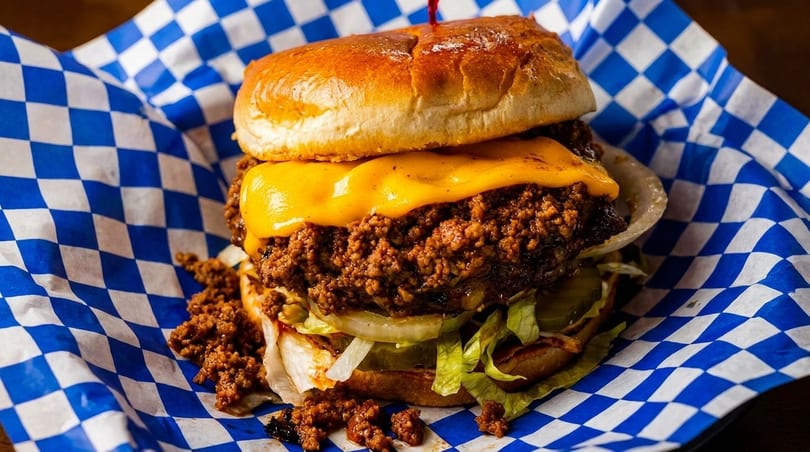 A sloppy joe with melted cheese, onions, lettuce, and pickles, served in blue and white checkered paper.