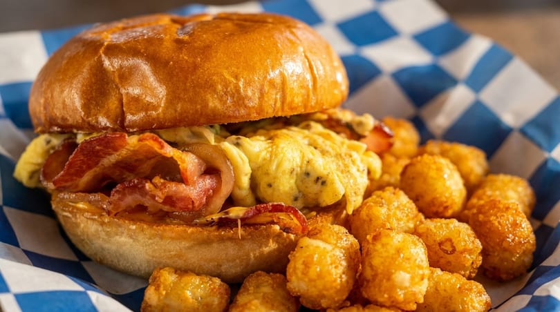 A breakfast sandwich with bacon and scrambled eggs, served with crispy tater tots in a checkered basket.